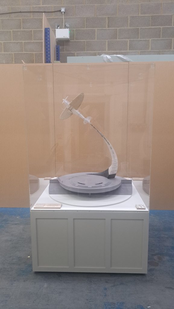 A display case contains a model of an aeroplane on a curved support above a circular base with markings, set against an industrial background with brick walls and blue metal shelving.