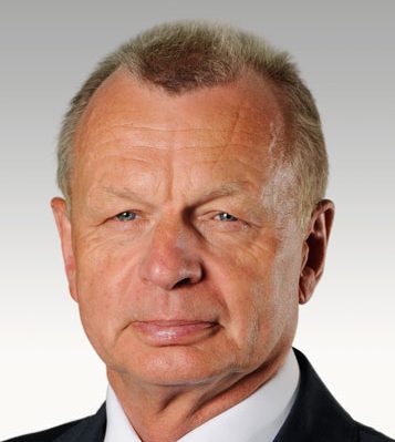 A middle-aged man with short, fair hair and blue eyes, wearing a dark suit and white shirt, is facing the camera with a neutral expression against a plain, gradient background.