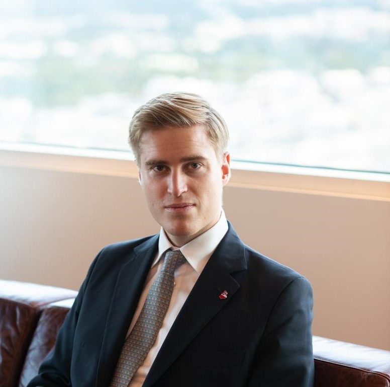 A man in a suit and tie sits on a brown leather sofa in front of a large window with a blurred cityscape in the background. He looks directly at the camera with a neutral expression.
