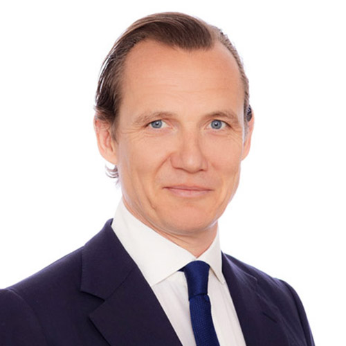 A man with light skin, blue eyes, and short, slicked-back brown hair is wearing a dark suit, white shirt, and dark tie, posing against a plain white background.