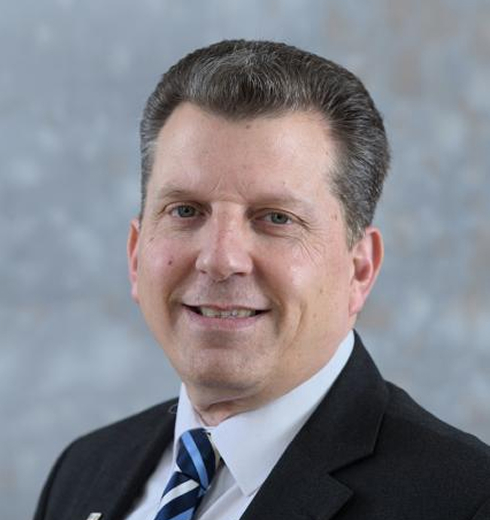 A man in a dark suit, white shirt, and striped tie smiles at the camera against a blurred grey background. He has short brown hair and fair skin.