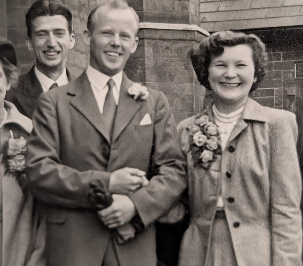 Three people dressed in formal attire, likely at a wedding. The man in front wears a suit with a buttonhole and holds the arm of a smiling woman wearing a corsage. Another man smiles in the background. The photo is black and white.