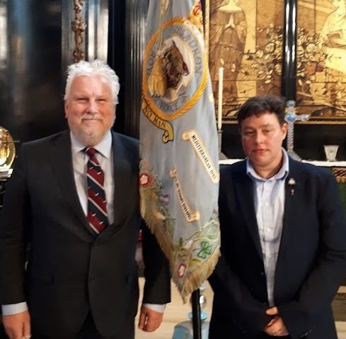 Two people in suits stand indoors beside a decorative flag with an emblem and text. The background features ornate decor, including a detailed tapestry and candle holders.