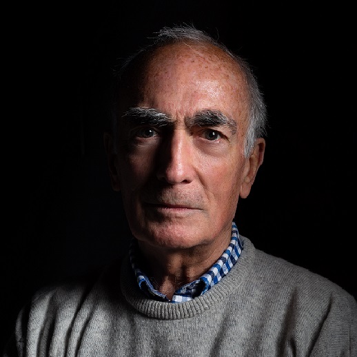 An older man with grey hair and thick eyebrows, wearing a grey jumper over a blue and white checked shirt, looks directly at the camera against a dark background.