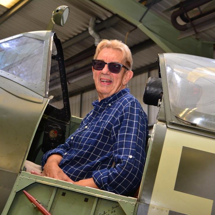 A person wearing sunglasses and a blue checked shirt smiles whilst sitting in the cockpit of a vintage aeroplane inside a hangar.