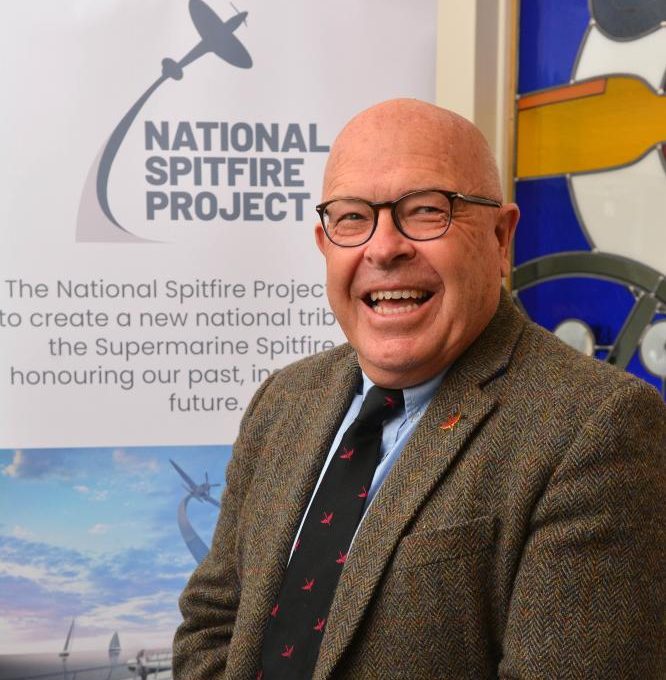 A smiling man wearing glasses, a brown tweed jacket, and a patterned tie stands in front of a National Spitfire Project banner and a colourful mural featuring a yellow aeroplane.