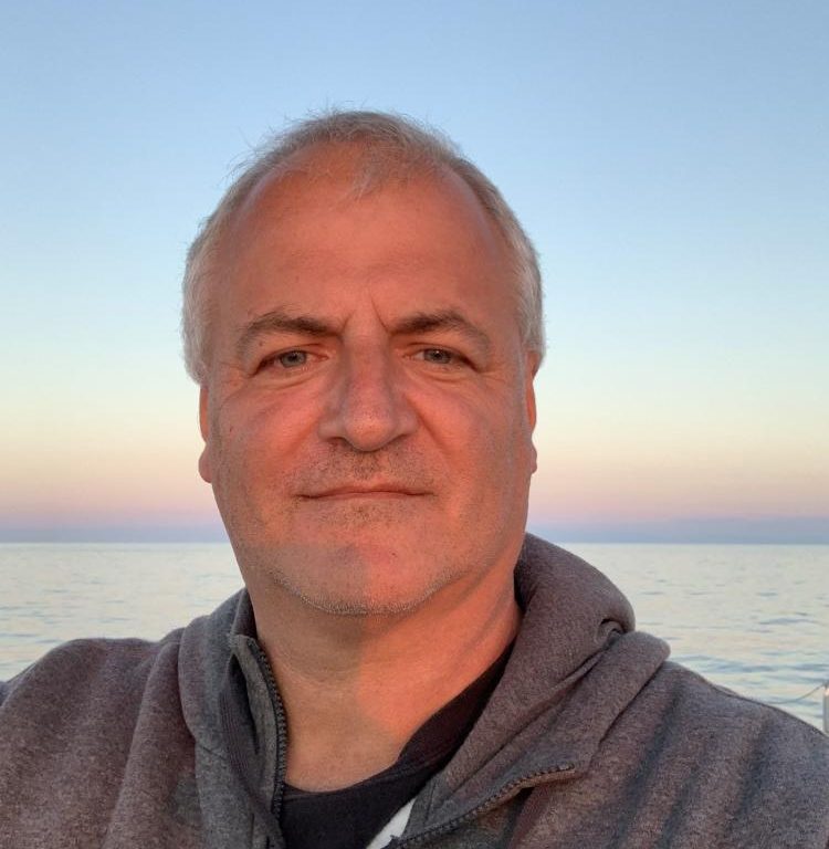 A middle-aged man with short grey hair and a slight smile takes a selfie outdoors by calm water at sunset, with a clear sky and soft pastel colours in the background. He is wearing a grey hoodie.