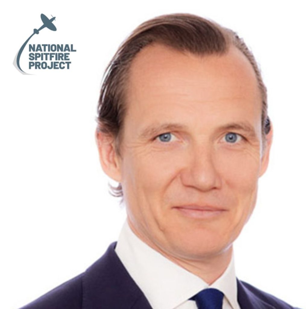 A man in a suit and tie with slicked-back hair is smiling slightly. The logo for the National Spitfire Project, featuring a silhouette of a Spitfire aeroplane, is in the top left corner on a white background.