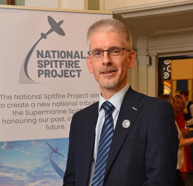 A man in a suit and striped tie stands smiling in front of a National Spitfire Project banner at an indoor event. The banner features a silhouette of a Spitfire aeroplane and descriptive text about the project.