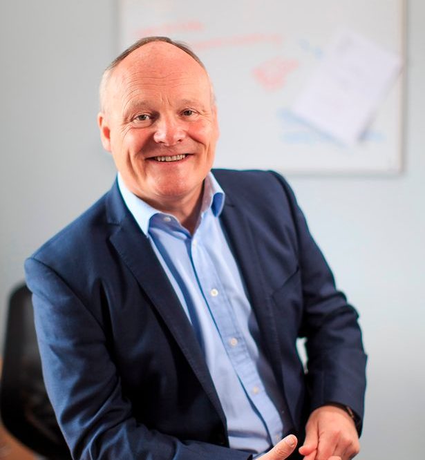 A smiling middle-aged man in a blue suit and light blue shirt sits indoors, in front of a blurred whiteboard with some writing and papers pinned to it.