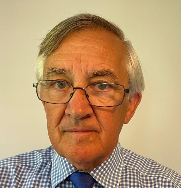 An older man with grey hair, wearing glasses, a blue checked shirt, and a blue tie, looks directly at the camera against a plain light-coloured background.