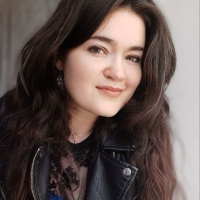 A young woman with long dark hair smiles softly at the camera. She is wearing a black leather jacket and a dark lace top, standing against a plain, light background.